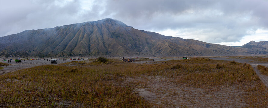Panoramic view of Bromo Tengger Semeru national park landscape with volcanic hills and green savanna under cloudy sky.