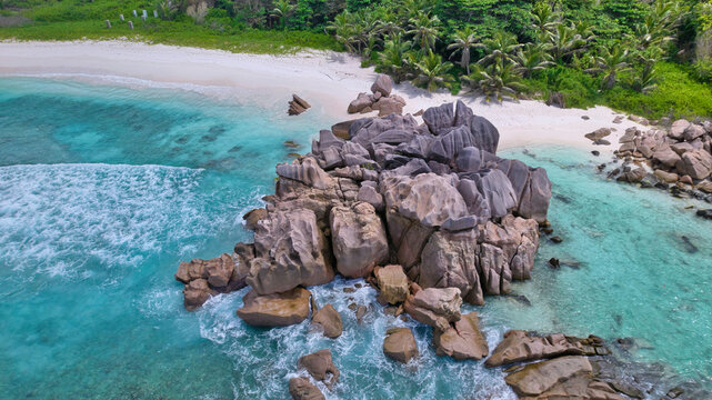 High-angle drone shot of dramatic granite boulders, white sandy beach, and vibrant tropical forest meeting the turquoise ocean at Anse Cocos, La Digue, Seychelles. Beautiful nature background