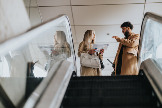 Two business colleagues discuss travel documents while standing on an escalator. They converse and gesture in a modern airport terminal as they prepare to board.