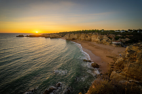 View of the golden sunset over the ocean waves hitting a sandy beach framed by rugged limestone cliffs and coastal vegetation in Albufeira, Faro District, Portugal.