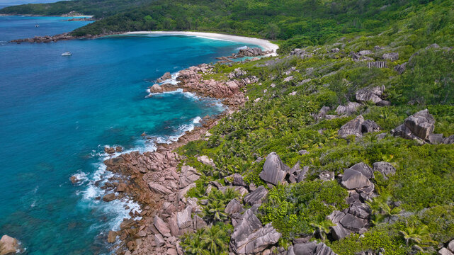 Breathtaking drone shot capturing the untamed beauty of Anse Cocos in Seychelles, showcasing clear turquoise ocean waves crashing against granite boulders and a remote white sand bay.
