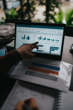 A business professional points at a laptop screen displaying analytics charts and graphs. The scene shows a close-up view of data visualization and printed business plan documents on a desk.