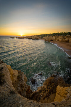 View of the sun setting over the golden cliffs and turquoise waters of a secluded beach Albufeira, Faro District, Portugal.