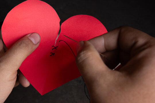 Close up of hands sewing a torn heart-shaped paper together, representing forgiveness, reconciliation, healing emotional wounds, rebuilding relationships, and starting over concept.