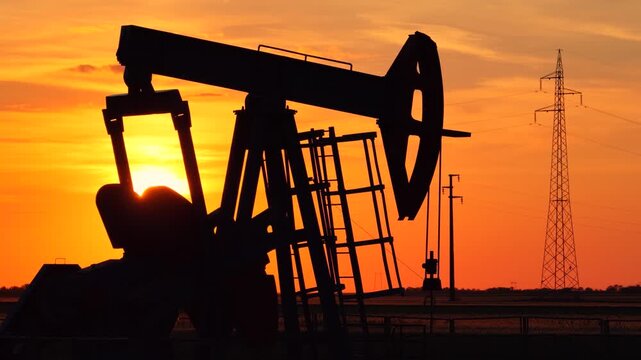 Silhouette of oil pump jack against a colorful beautiful sky with glowing clouds at sunset, high voltage electric pole in background