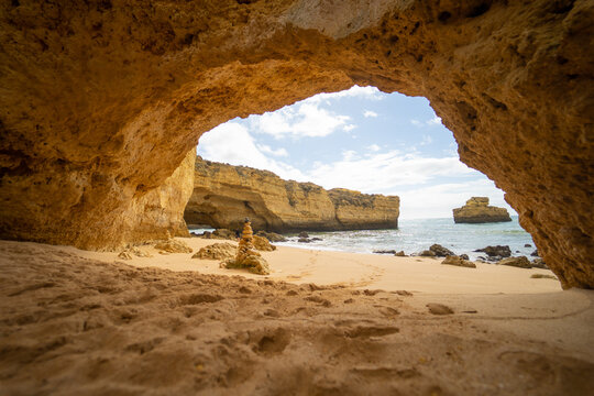 View of a sandy beach and turquoise ocean through a natural rock archway with golden limestone cliffs and a stone stack in Albufeira, Faro District, Portugal.
