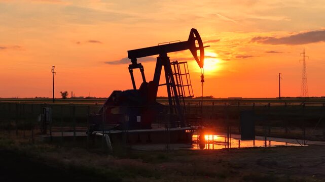 Silhouette of oil pump jack against a colorful beautiful sky with glowing clouds at sunset, high voltage electric pole in background