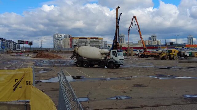 Cement mixer trucks move around an urban construction site. Workers are busy preparing the land for new buildings. Cranes lift materials high into the air. The site is located in a city environment wi