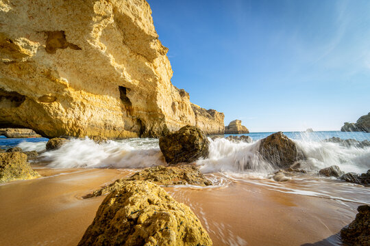 View of waves crashing against golden rocks and limestone cliffs on a sandy beach under a clear blue sky in Albufeira, Faro District, Portugal.