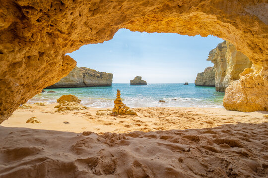 View of a sandy beach with a stone stack through a limestone cave opening under a clear blue sky in Albufeira, Faro District, Portugal.