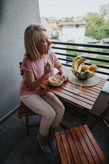 Obraz premium A middle-aged woman sits on a small balcony eating a bowl of salad. She has a light meal at a wooden table beside a bowl of fruit overlooking the neighborhood.