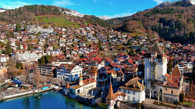 4K aerial drone footage over Oberhofen am Thunersee, Switzerland - Oberhofen Castle on Lake Thun, lakeside village with Swiss chalets, snow-capped Alps, and Bernese Oberland in winter.