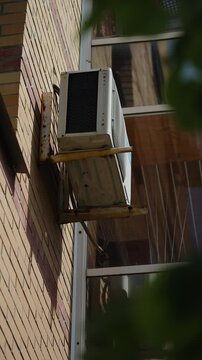 Exterior air conditioner unit mounted on a brick apartment wall, secured by rusted metal brackets beside a glazed balcony window, photographed from below in soft summer light