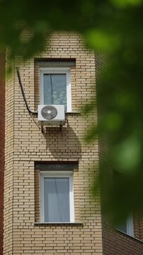 Vertical view of a light brick apartment facade with two white-framed windows and a wall-mounted air conditioner, framed by green foliage in the foreground on a sunny urban day