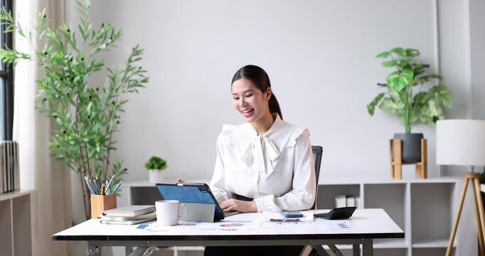 Young asian business manager calculating expenses on a desktop calculator and looking at a tablet, managing financial data while conducting her work in a contemporary office setting