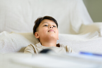 Tired Asian preteen boy resting his head on a comfortable sofa after finishing homework. Representing study breaks, childhood relaxation, daydreaming, and domestic learning routines © Kornkanok
