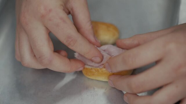 Hands place ham slices onto a small bread roll for a sandwich
