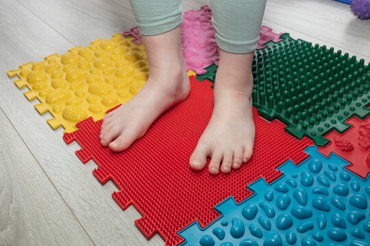 Child standing barefoot on a red textured sensory massage mat. Concept of sensory integration, balance development, physical therapy, wellness, foot health.