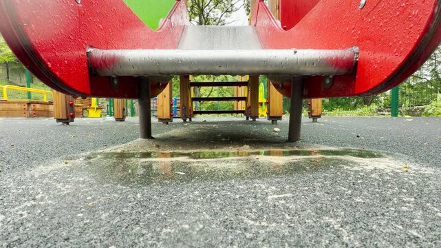 rainwater drips off the edge of a playground structure. The falling water makes a soft plop sound as it hits the puddle below. This scene brings an empty, melancholic feeling to the quiet park.