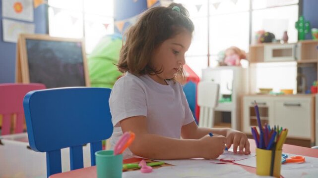 Girl drawing with pencil at preschool classroom table, leaning on elbow and tracing paper; concentration creativity learning.