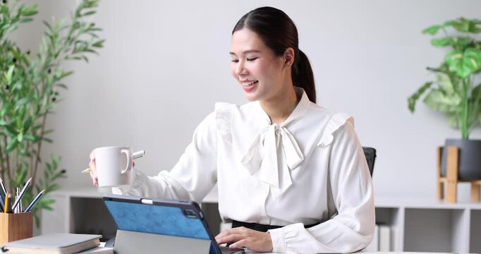 Young asian business manager calculating expenses on a desktop calculator and looking at a tablet, managing financial data while conducting her work in a contemporary office setting