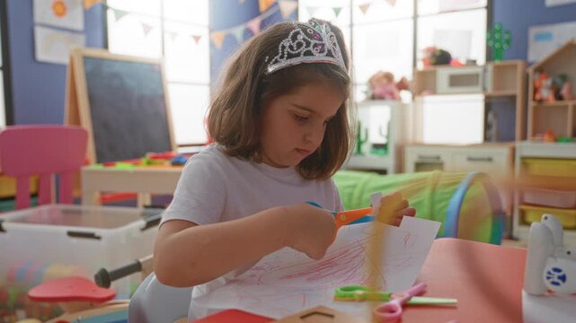 Girl cutting paper with child scissors at a colorful preschool classroom table, hands holding scissors and drawing sheet, wearing a plastic tiara and white shirt; concentration.