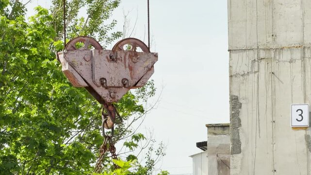 A massive metal pulley and hook sway gently in the air next to a concrete wall. The heavy machinery operates continuously on the development site. This active work creates a loud industrial hum.