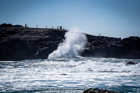 View of powerful waves crashing against dark volcanic cliffs with sea spray and people standing on the ridge under a clear blue sky in El Cotillo, Canary Islands, Spain.