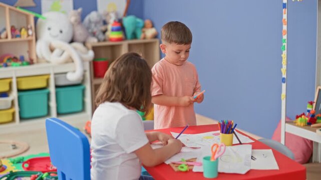 Boy holds orange scissors and girl draws on paper at a building playroom table with colored pencils and crafts; playful friendship.