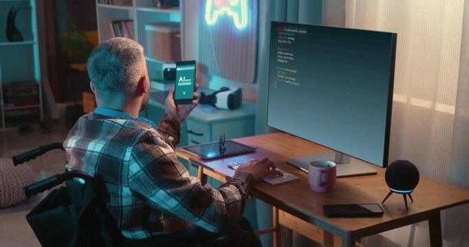 Man in wheelchair uses an AI assistant on his smartphone while programming at a computer in a home office. Artificial intelligence supporting everyday work and productivity.