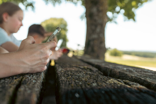 Three children sitting at a wooden picnic table on a phone screen, view through tree trunks, conceptual shot for digital dependency