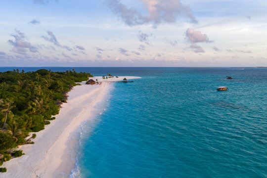 Aerial view of a narrow sandspit curving from a palm island in the Maldives, with beach huts, loungers, and boats on a lagoon in warm evening light and soft clouds.