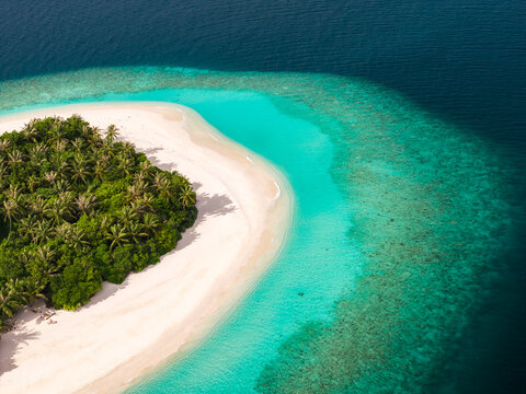 Aerial view of a Maldives coral atoll with a white sand crescent, palm trees, and a turquoise lagoon fading to deep sapphire, calm seas, and a clear reef edge.