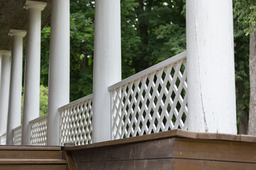 Naklejka premium A peaceful exterior view of a classic cottage porch featuring tall white columns, diamond-pattern lattice railing and weathered wooden steps framed by green summer trees and soft natural light