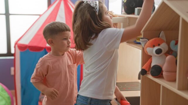Girl reaching for toy on shelf inside a playroom building as boy watches nearby, plush fox and toy stove visible; playful joy friendship.