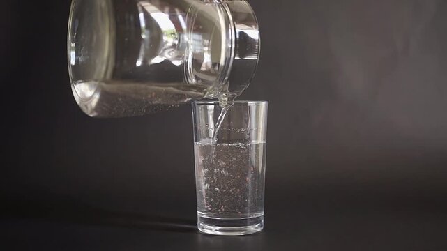 Hand pouring drinking water from a glass pitcher into a drinking glass on a black background, illustrating the concept of microplastic contamination