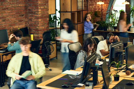Busy creative office with employees working and moving between desks with motion blur. Concept of workplace productivity, agency workflow, multitasking, digital business, office operations.