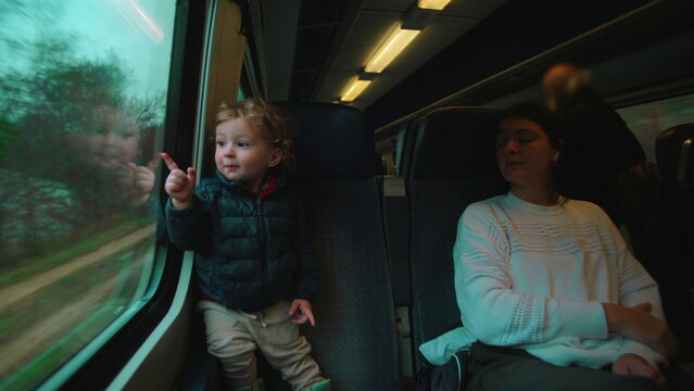 Toddler standing on train seat pointing at passing countryside through window while parent rests nearby evoking curiosity shared travel bonding attention early memory and quiet discovery