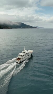 Aerial Drone Tracking Shot White Luxury Yacht Speeding on Ocean with Mountain and Cloudy Sky Background