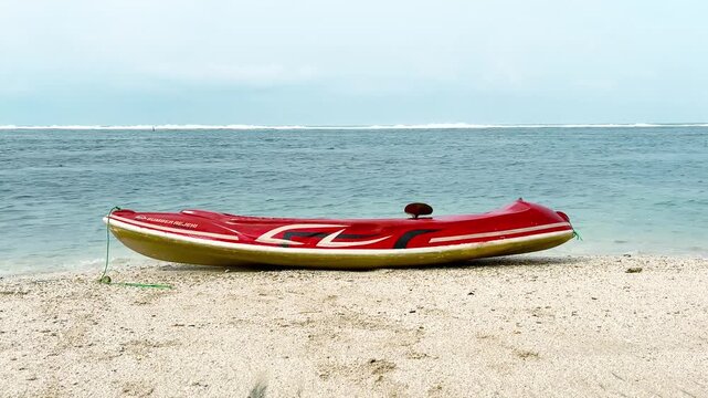 Garut, West Java, Indonesia - June 15, 2026; A red canoe sits on the shore. White sand and blue sea water create a beautiful, sunny day. A beautiful beach.