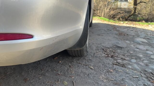 Rear wheel rolls over uneven gravel as a silver car moves forward, close-up of tire rotation, suspension response, rough road travel, automotive motion.