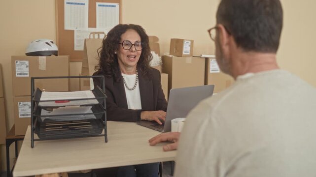 Woman shakes hand over stacked boxes and a laptop at a packing desk in a small business building; trust partnership agreement.