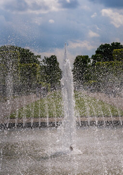 Dynamic water jets from a fountain creating a misty spray against a backdrop of lush green trees and a cloudy sky at Herrenhausen Gardens.