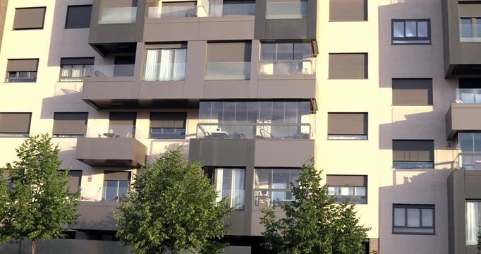 Close view of a modern residential apartment building featuring glass balconies, large windows and contemporary facade design. Urban housing architecture with trees in the foreground in Madrid, Spain.