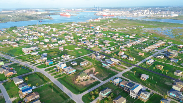 Industrial horizon coastal mix presents elevated homes arranged in tight Surfside grid, harbor channel, cranes, anchored vessels stretching across background. Seaside district in Freeport, TX