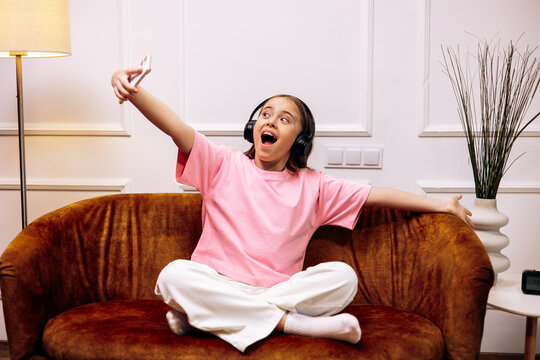 Young girl wearing headphones and pink t-shirt taking a selfie, singing along to music while relaxing on a couch at home