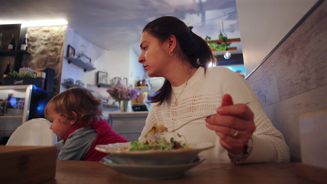 Mother attempting to eat at restaurant while holding restless toddler on lap conveying multitasking stress divided attention and everyday challenges of parenting in public space