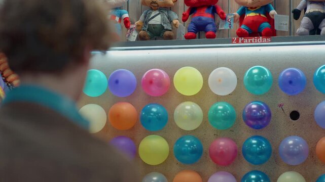 Colorful balloons pinned to wall in dart popping game at fairground arcade, plush toy prizes above, dart with flag thrown at target, missing the shot