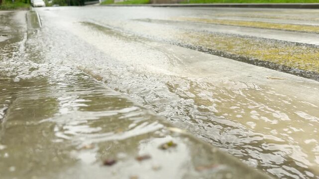 heavy rain sends streams of water rushing down the concrete street gutter. The moving current creates steady bubbling and sloshing sound. Tires rolling through the wet crosswalk add a swishing noise.