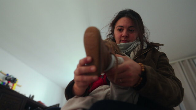 Mother helping toddler put on shoe indoors preparing for outing supporting independence daily routine caregiving moment expressing patience protection and family bond